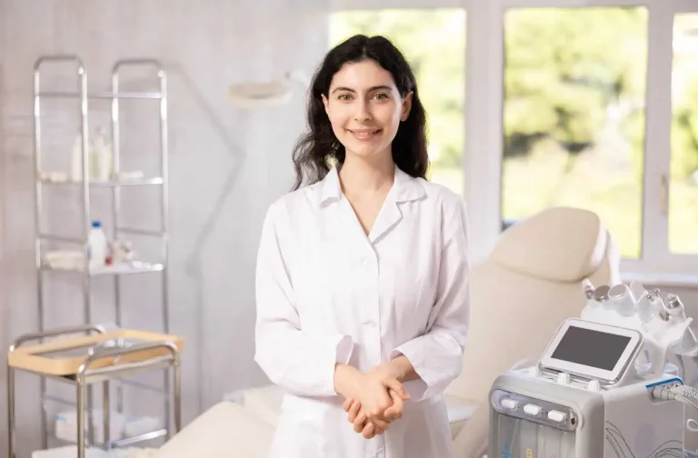 A med spa practitioner in a white coat standing beside her treatment chair and equipment